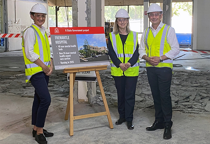 Two women and man wearing hi visibility safety vests, goggles and hard hats stand in a construction zone in front of a poster on an easel. The poster reads Fremantle Hospital, 40 new mental health beds, new 24-hour mental health urgent care service, opening in 2025